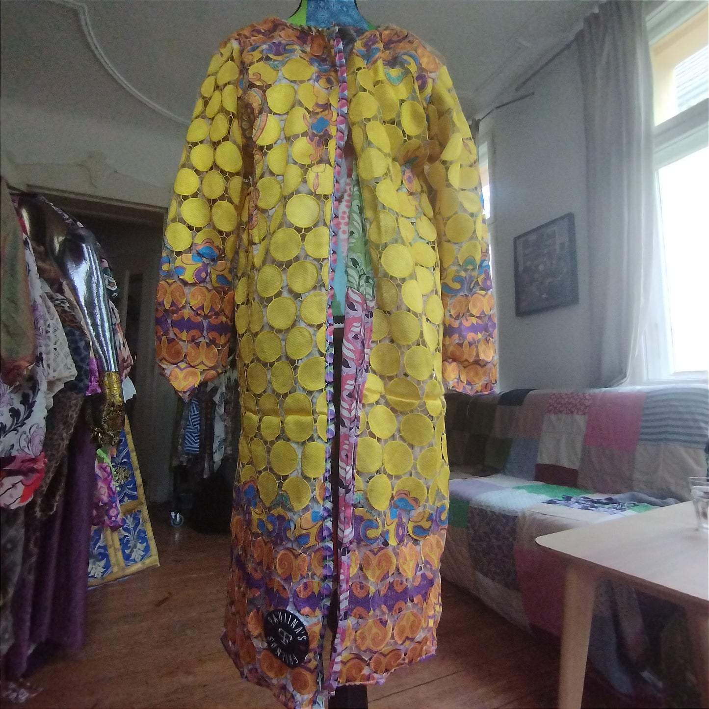 Close-up view of a vibrant, intricately designed fabric detail from an upcycled vintage kimono-style coat. Features bold yellow lace patterns, colorful floral embroidery, and a mix of rich, festive prints inspired by Canarian carnival aesthetics. A striking combination of textures and colors for a unique, eye-catching garment.
