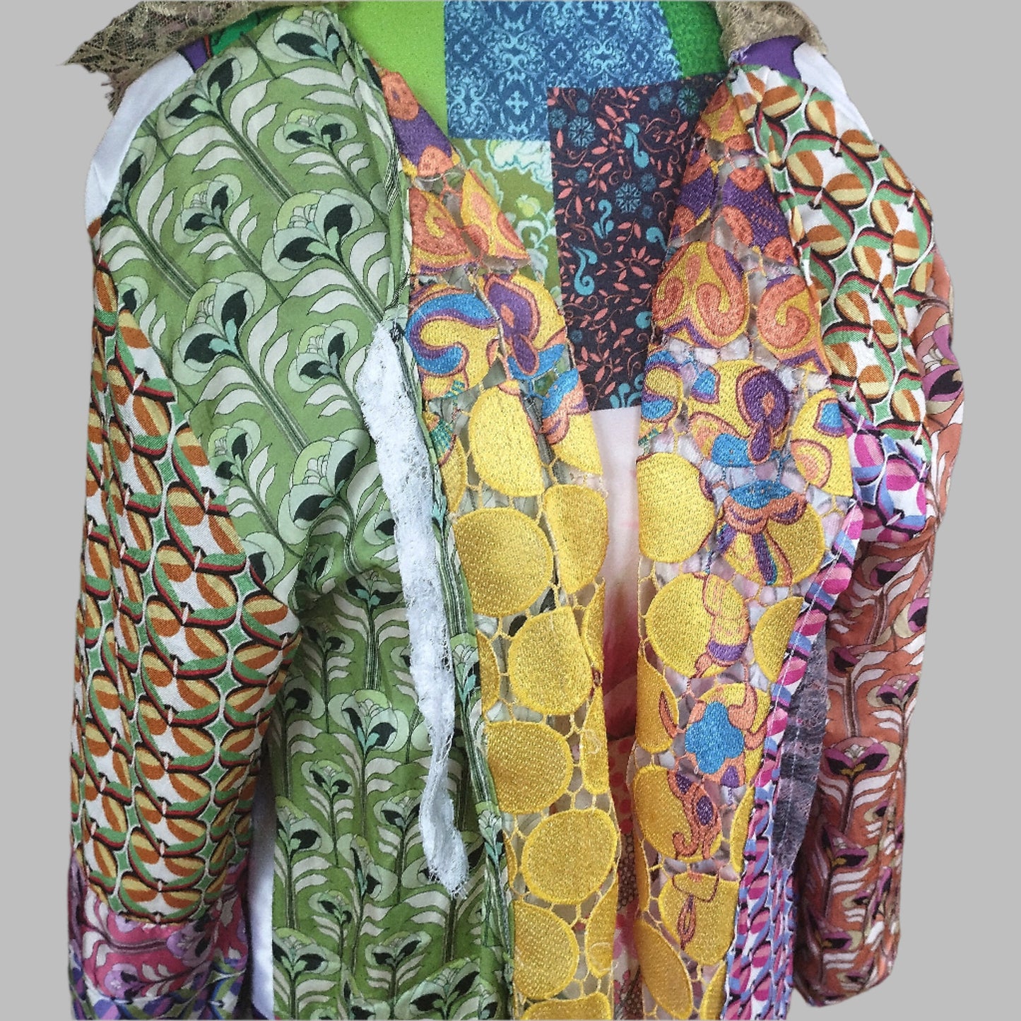 Close-up view of a vibrant, intricately designed fabric detail from an upcycled vintage kimono-style coat. Features bold yellow lace patterns, colorful floral embroidery, and a mix of rich, festive prints inspired by Canarian carnival aesthetics. A striking combination of textures and colors for a unique, eye-catching garment.