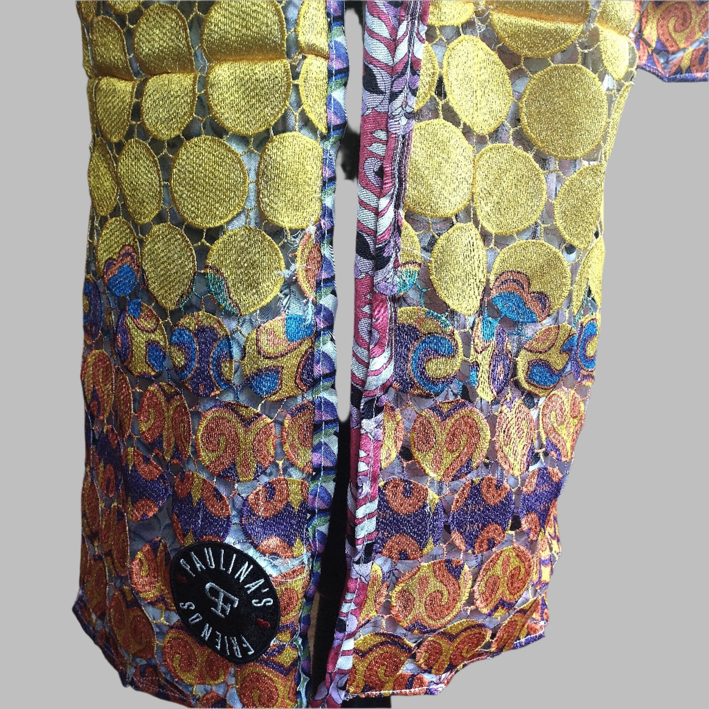 Close-up view of a vibrant, intricately designed fabric detail from an upcycled vintage kimono-style coat. Features bold yellow lace patterns, colorful floral embroidery, and a mix of rich, festive prints inspired by Canarian carnival aesthetics. A striking combination of textures and colors for a unique, eye-catching garment.