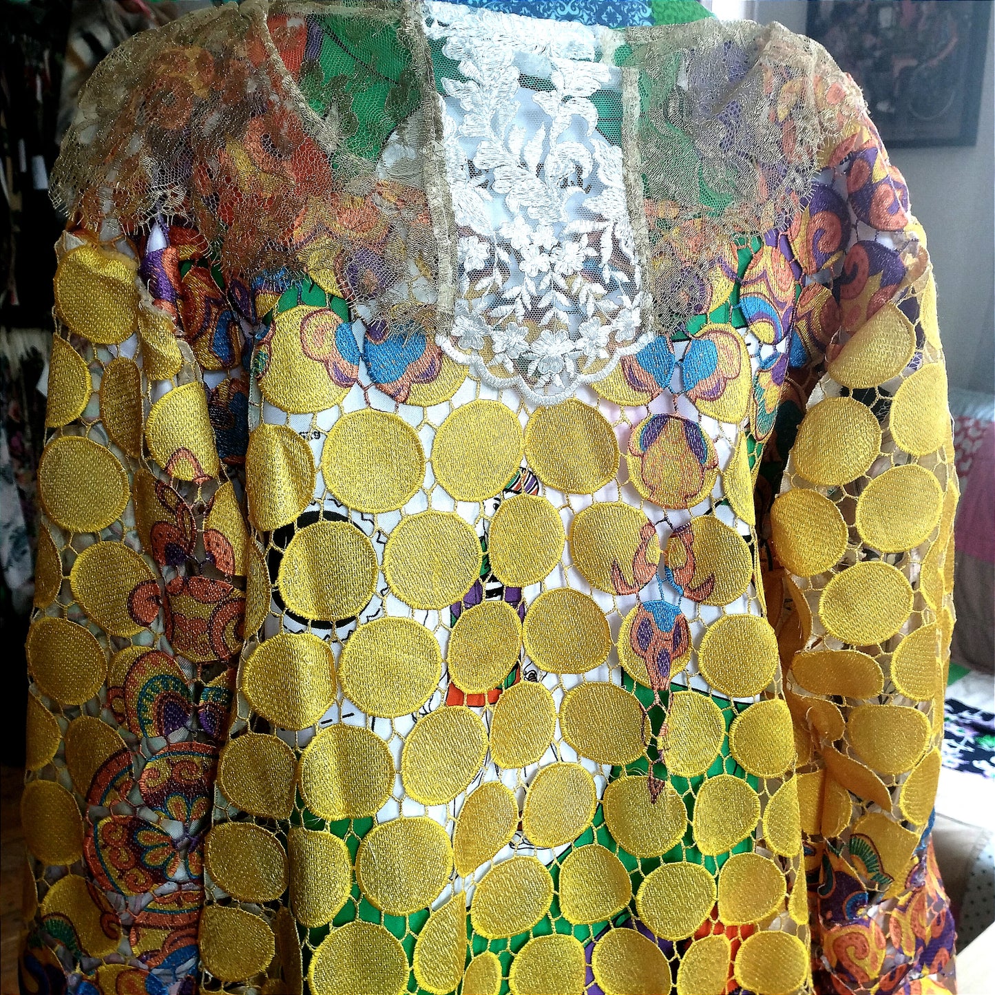 Close-up view of a vibrant, intricately designed fabric detail from an upcycled vintage kimono-style coat. Features bold yellow lace patterns, colorful floral embroidery, and a mix of rich, festive prints inspired by Canarian carnival aesthetics. A striking combination of textures and colors for a unique, eye-catching garment.