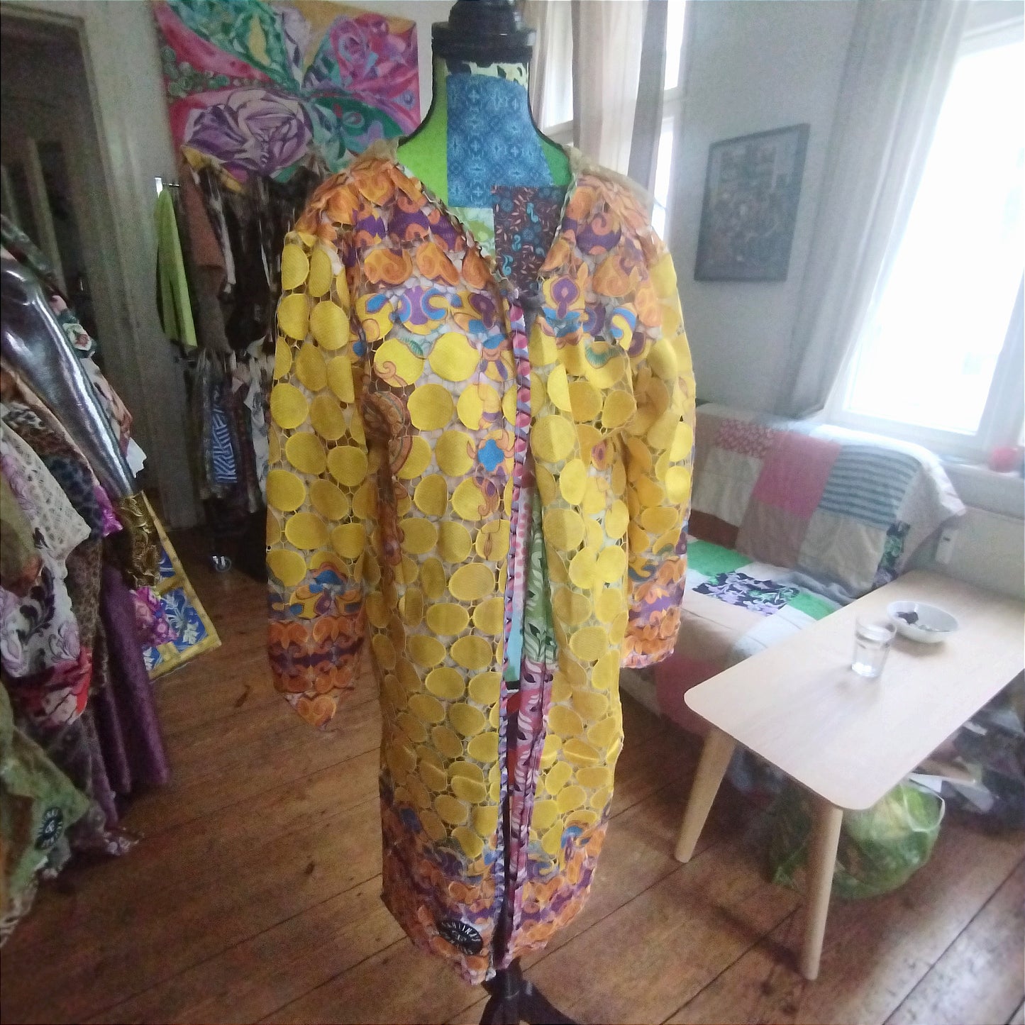 Close-up view of a vibrant, intricately designed fabric detail from an upcycled vintage kimono-style coat. Features bold yellow lace patterns, colorful floral embroidery, and a mix of rich, festive prints inspired by Canarian carnival aesthetics. A striking combination of textures and colors for a unique, eye-catching garment.