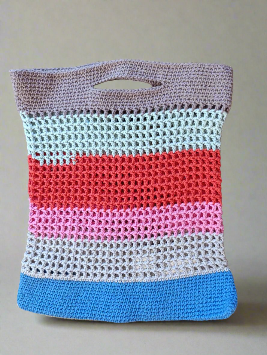 Hand-crocheted tote bag with multicolored horizontal stripes in shades of beige, blue, pink, and red, featuring an integrated handle and open-weave design.