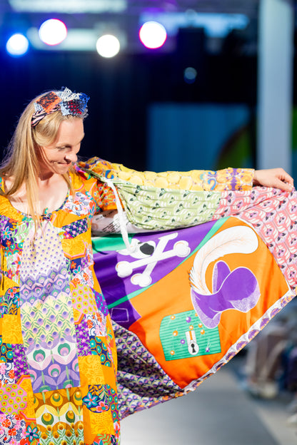 Close-up view of a vibrant, intricately designed fabric detail from an upcycled vintage kimono-style coat. Features bold yellow lace patterns, colorful floral embroidery, and a mix of rich, festive prints inspired by Canarian carnival aesthetics. A striking combination of textures and colors for a unique, eye-catching garment.