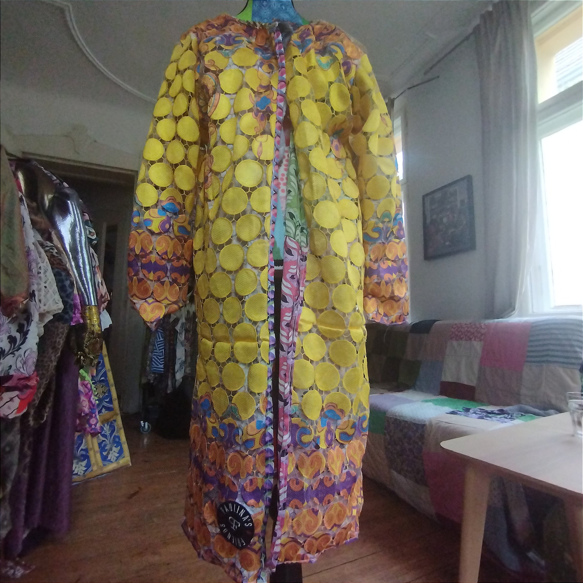 Close-up view of a vibrant, intricately designed fabric detail from an upcycled vintage kimono-style coat. Features bold yellow lace patterns, colorful floral embroidery, and a mix of rich, festive prints inspired by Canarian carnival aesthetics. A striking combination of textures and colors for a unique, eye-catching garment.
