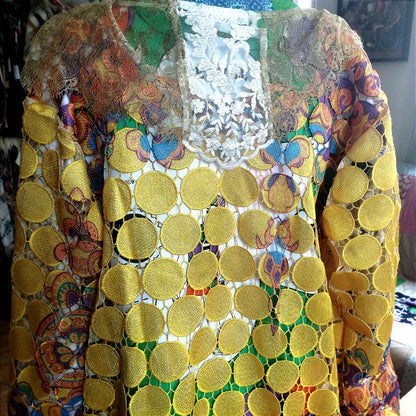 Close-up view of a vibrant, intricately designed fabric detail from an upcycled vintage kimono-style coat. Features bold yellow lace patterns, colorful floral embroidery, and a mix of rich, festive prints inspired by Canarian carnival aesthetics. A striking combination of textures and colors for a unique, eye-catching garment.