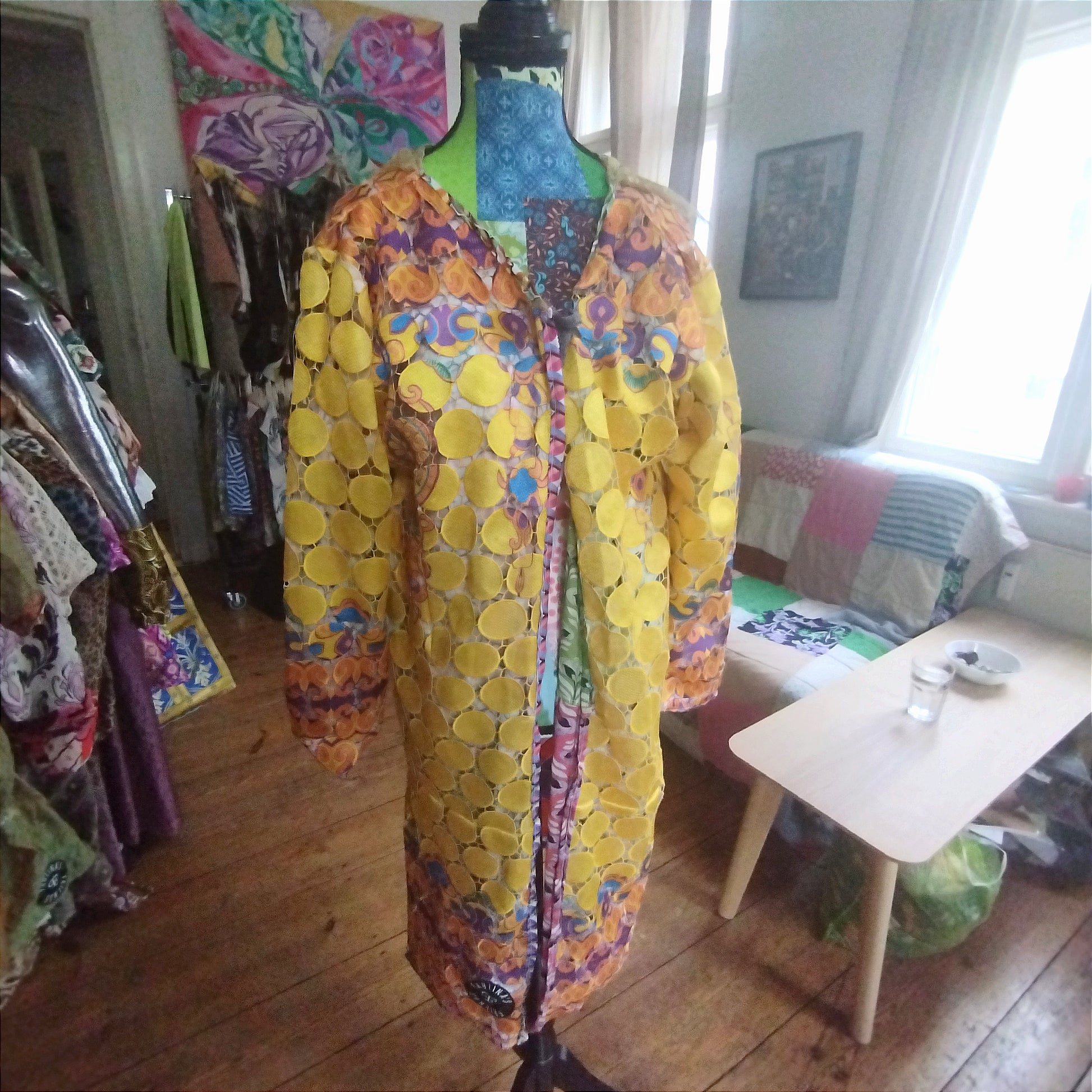 Close-up view of a vibrant, intricately designed fabric detail from an upcycled vintage kimono-style coat. Features bold yellow lace patterns, colorful floral embroidery, and a mix of rich, festive prints inspired by Canarian carnival aesthetics. A striking combination of textures and colors for a unique, eye-catching garment.